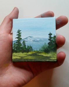 a hand holding a small piece of art with a mountain in the background and trees