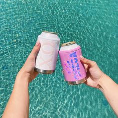 two people holding up pink and white cans next to blue water with their hands in the air