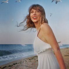 a woman in a white dress standing on the beach with birds flying above her head