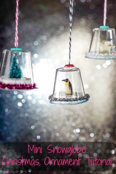 three glass bell ornaments hanging from strings in front of glittery background with penguin and snowflakes