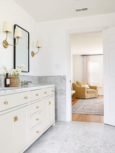 a white bathroom with marble counter tops and gold hardware on the vanity, along with a beige chair