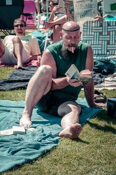 an older man sitting on the grass reading a book while others sit in the background