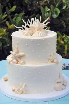 a three tiered white cake with sea shells and starfish on the top, sitting on a blue table cloth