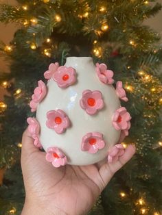 a hand holding a white vase with pink flowers on it next to a christmas tree