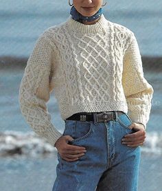 a woman standing on the beach with her hands in her pockets wearing a sweater and jeans