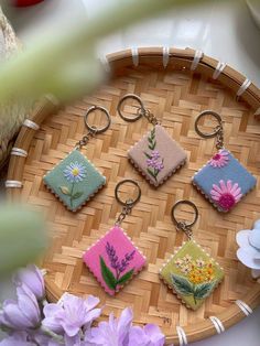 six needle - embroidered keychains are arranged on a woven tray with purple flowers