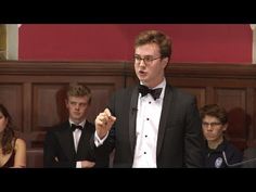 a young man in a tuxedo speaking at a podium with other people behind him