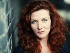 a woman with red hair and blue eyes looking at the camera while leaning against a brick wall