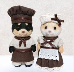 two small stuffed animals dressed in brown and white outfits, standing next to each other