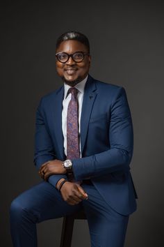 a man in a blue suit and tie sitting on a stool smiling at the camera