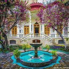 a fountain in front of a building with flowers on the ground and trees around it