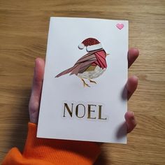 a person holding up a card with a bird on it and the word noel written in gold