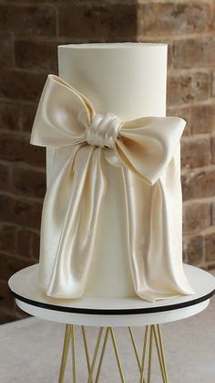 a white wedding cake with a bow on the top and gold wire stand in front of a brick wall