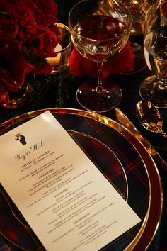 a menu sitting on top of a gold plate next to wine glasses and red roses