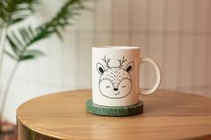 a coffee mug with a drawing of a deer on it sitting on a table next to a potted plant