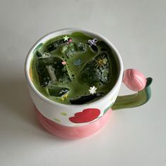 a cup filled with green liquid sitting on top of a white table next to a pink flower