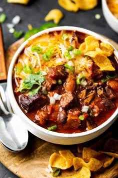 a white bowl filled with chili and tortilla chips on top of a wooden cutting board