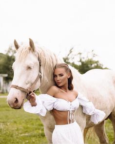 a beautiful woman standing next to a white horse in a field with her arm around the neck