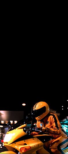 a man sitting on top of a yellow motorcycle at night with the lights on behind him