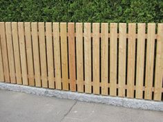 a wooden fence next to a sidewalk with bushes in the back ground and grass behind it