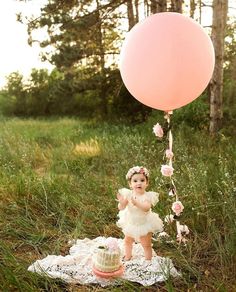 a baby girl in a white dress standing next to a pink cake and holding a balloon