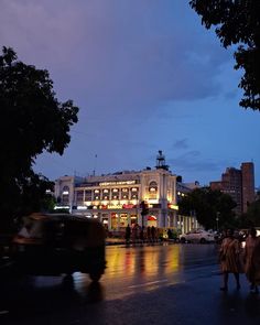 people are walking down the street at night in front of a large building with lights on it