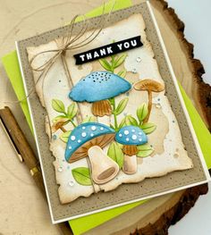 a thank you card with blue mushrooms and green leaves on the front, sitting on top of a piece of wood