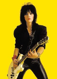 a woman with black hair holding an electric guitar and posing for a photo in front of a yellow background