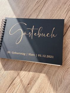 a black and gold wedding guest book sitting on top of a wooden table next to a pen