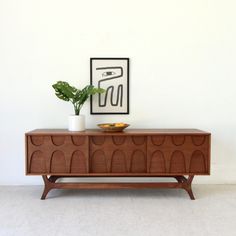 a wooden sideboard with a plant on top and an art piece above it in the corner