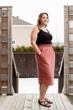 a woman standing on a wooden deck wearing a black tank top and pink midi skirt