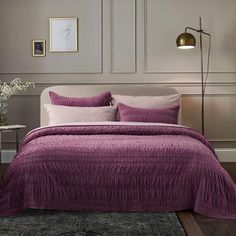 a bed with purple bedspread and pillows in a gray room next to a lamp