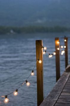 some lights that are on the side of a wooden pier near water with mountains in the background