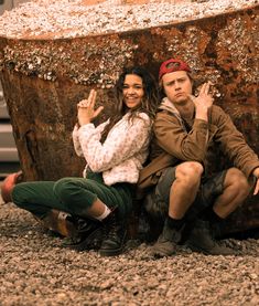 a man and woman sitting on the ground next to a tree stump with their hands in the air