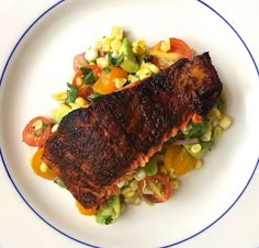 a white plate topped with grilled fish next to corn and tomato salad on top of it