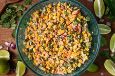 a glass bowl filled with corn salad on top of a wooden table next to limes