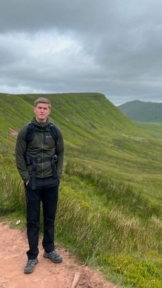 a man standing on top of a dirt road next to a lush green hill covered in grass