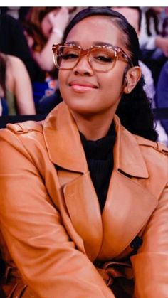 a woman sitting in front of a crowd wearing glasses and a leather jacket with her arms crossed