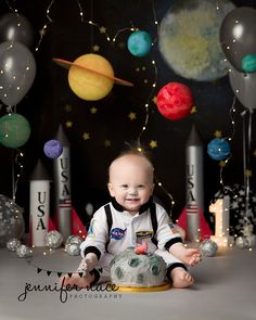 a baby is sitting on the floor in front of balloons and rockets, smiling at the camera