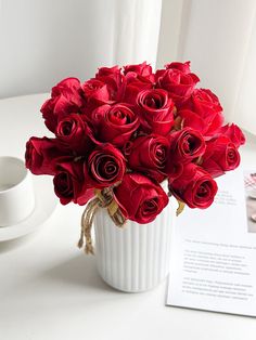 a white vase filled with red roses on top of a table next to a cup and saucer