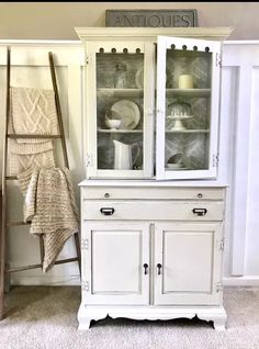 a white china cabinet with glass doors and drawers in a room next to a ladder