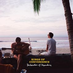 two men sitting on the beach playing guitar and singing to each other with an ocean in the background