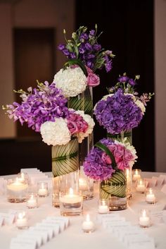 purple and white flowers are in vases on a table with lit candles around them