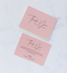 two pink wedding thank cards on a white marble surface with the word thank you written in cursive ink