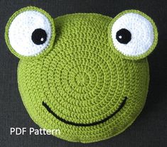 a crocheted frog pillow with eyes and nose on it's face, sitting on a black surface