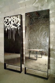 two mirrors with graffiti on them in front of a white brick wall, one has a bench