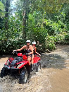 two people riding on four wheelers through muddy water in the jungle with trees and bushes behind them