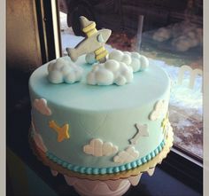 a blue cake with white clouds and an airplane on top is sitting in front of a window