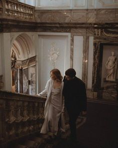 a man and woman are walking down the stairs in an ornate building with statues on either side