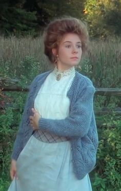 a woman in a dress and cardigan is standing by a fence with her arms crossed